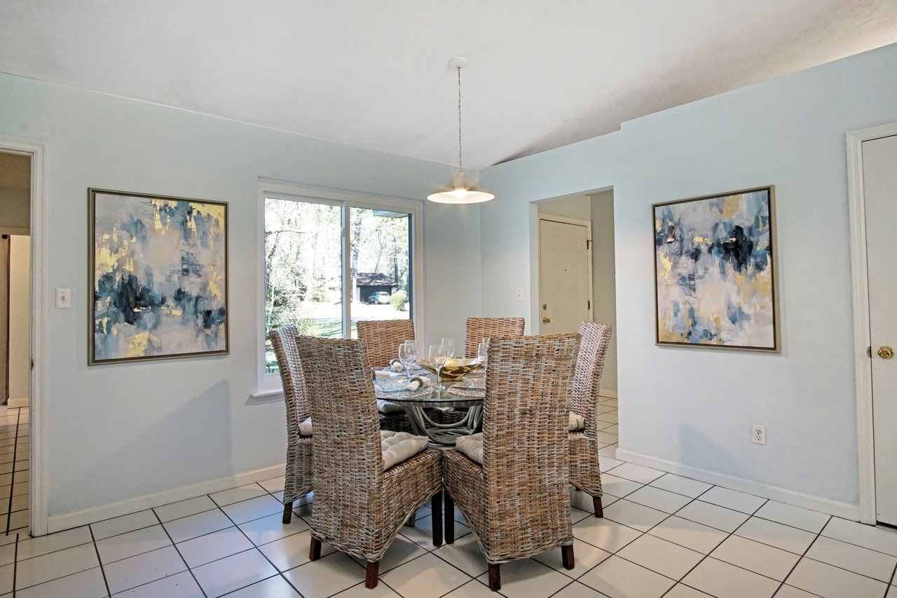 Bright dining room with a glass table surrounded by wicker chairs, large window showing greenery. Abstract paintings on pale blue walls. Airy and inviting.
