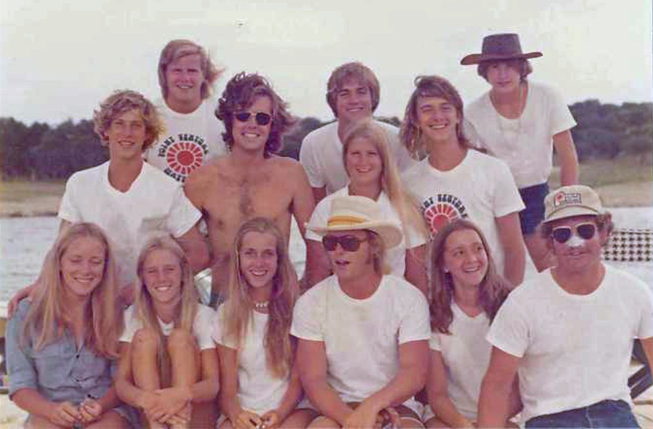 Thirteen young people pose outdoors beside the water, most wearing white shirts.
