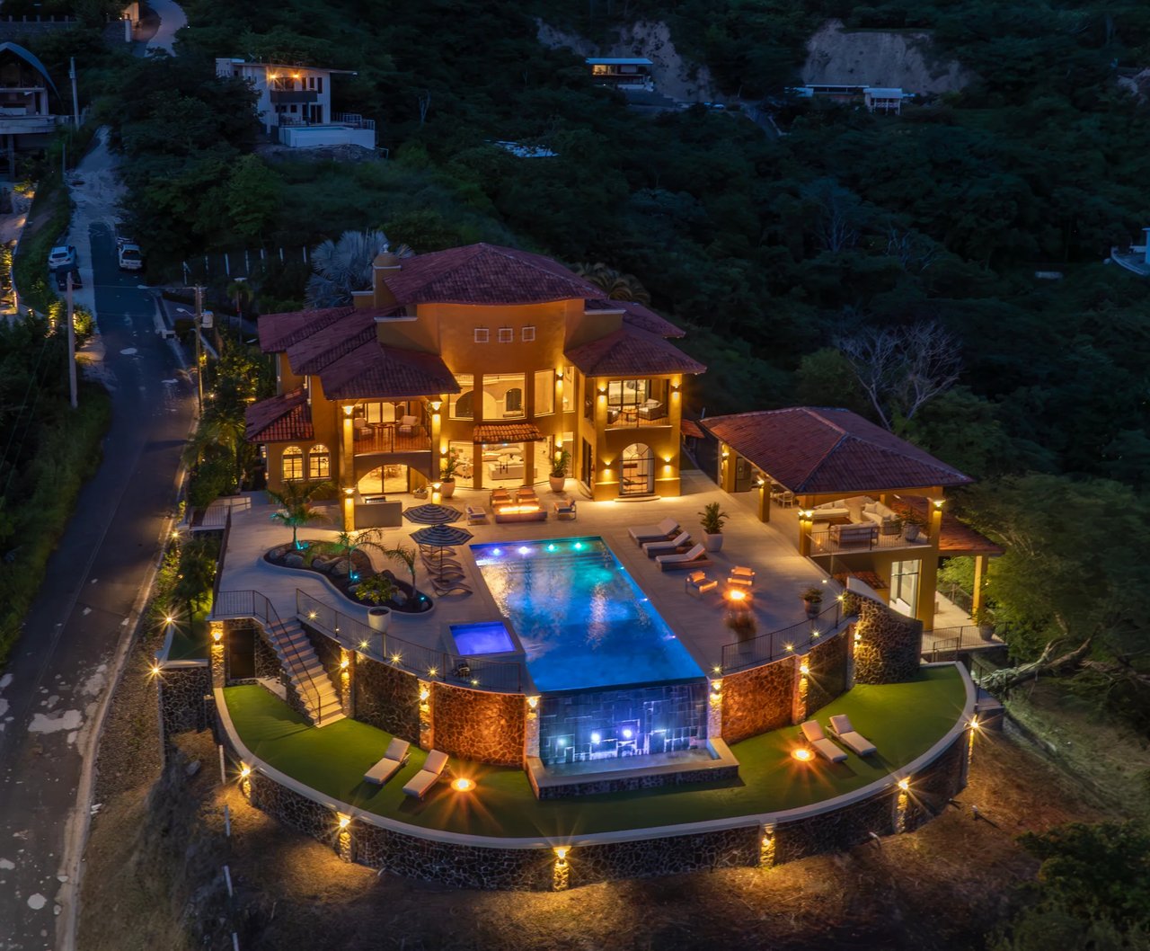 Aerial view of a lit luxury home with a swimming pool and forest.