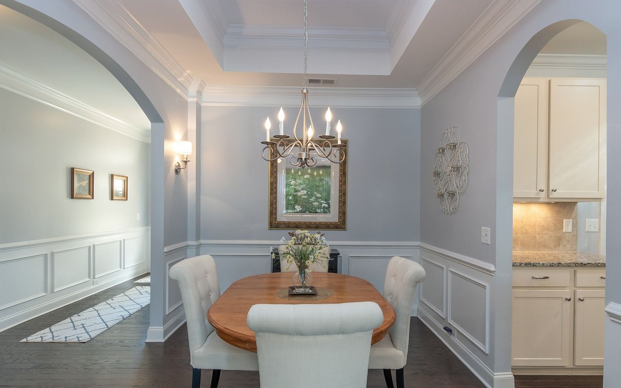 Dining room with a round wooden table, white upholstered chairs, and a chandelier beneath a tray ceiling.