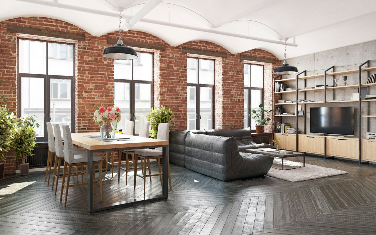 Loft-style living and dining room with exposed brick, tall windows, and a gray sectional sofa.