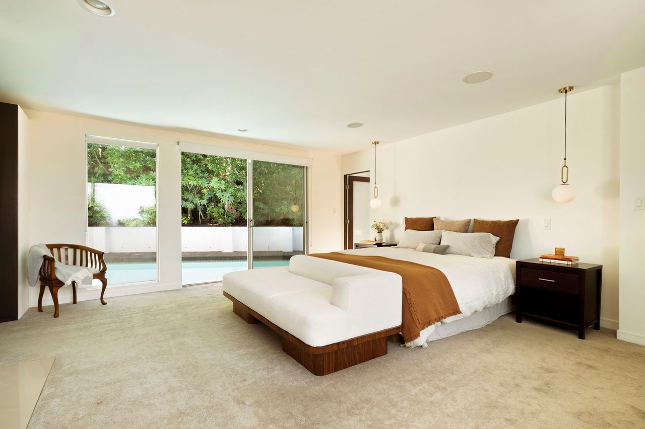 Bright bedroom with a king bed and bench beside sliding doors overlooking a pool and garden.