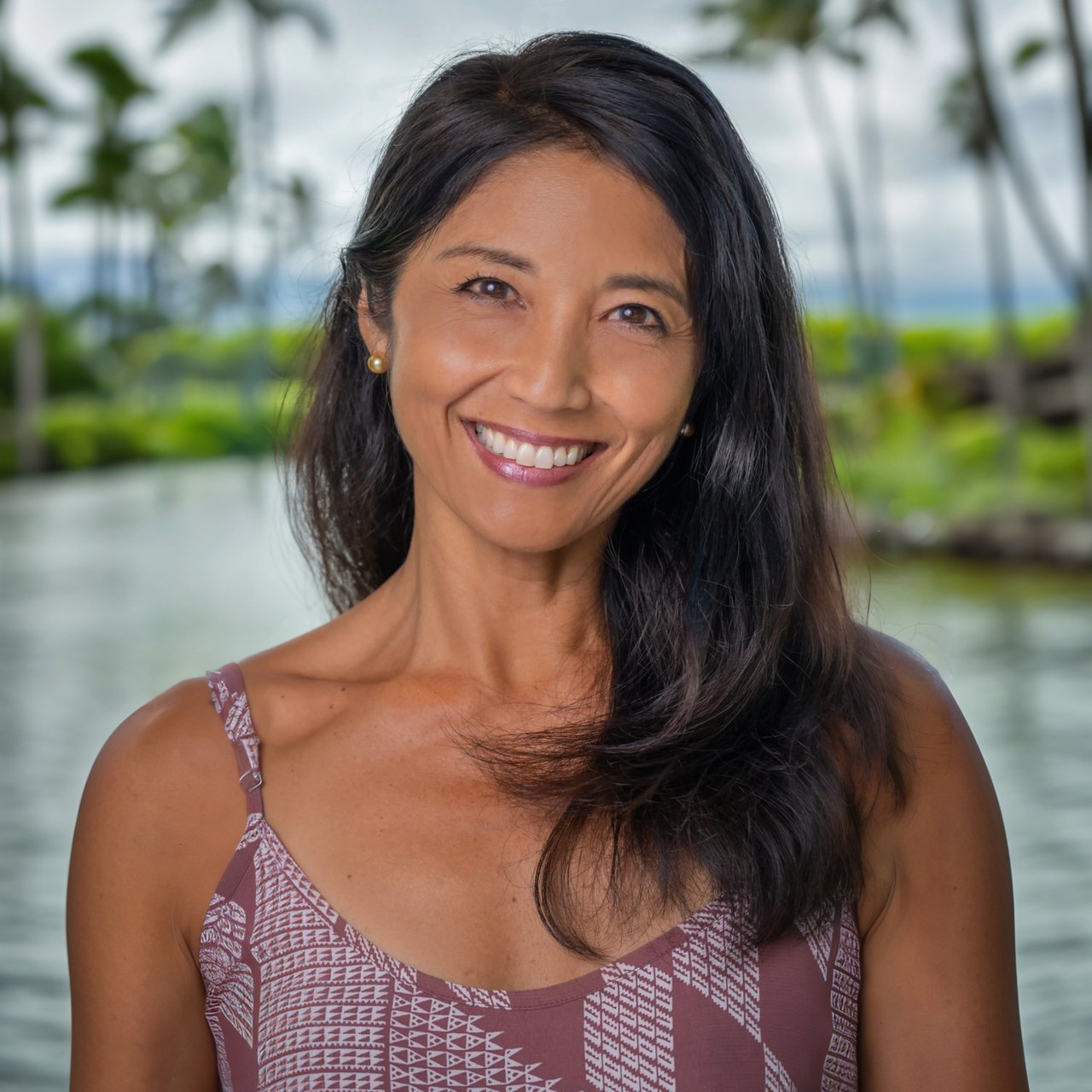 Debbie Edgerton smiling in a patterned mauve top beside tropical greenery and water.