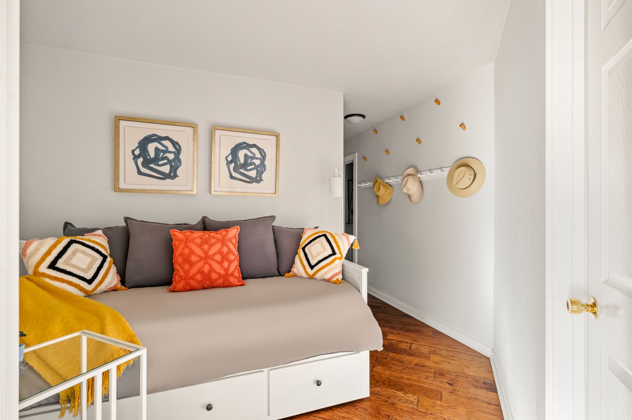 Staged bedroom with a built-in daybed, colorful pillows, framed artwork, and hats hanging on the wall.