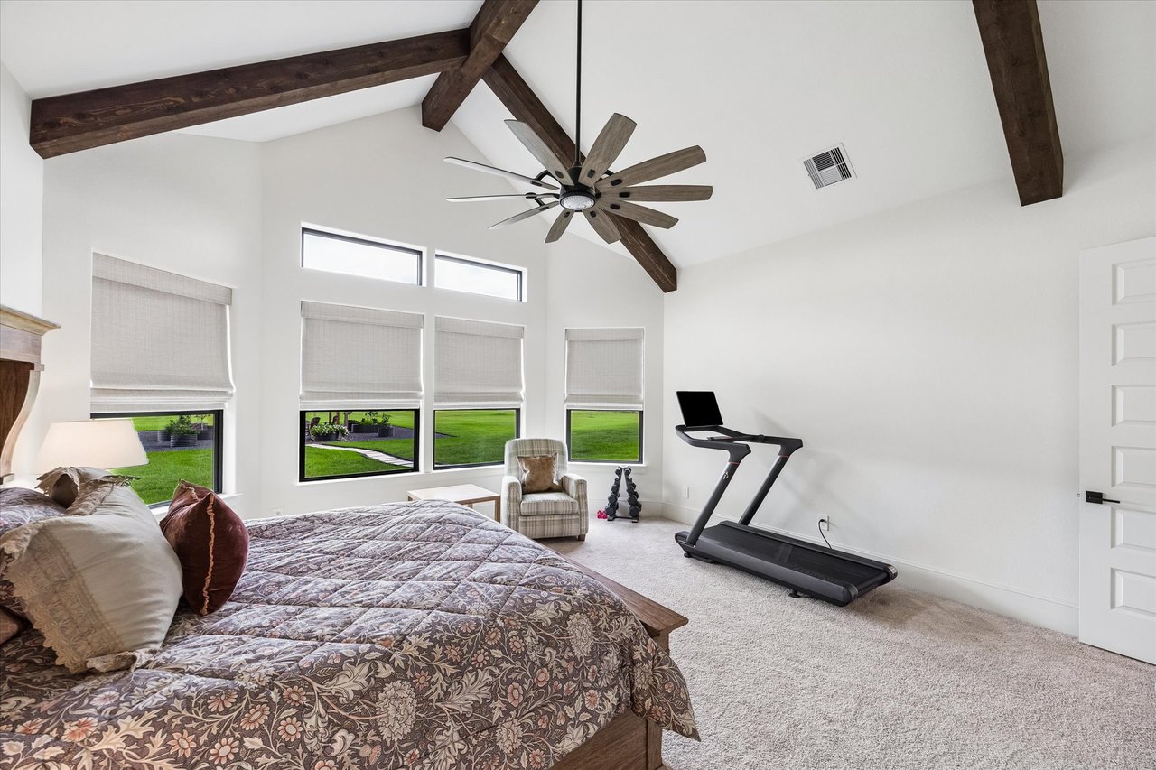 Spacious bedroom with vaulted wood-beamed ceiling, treadmill, and windows overlooking a lawn.