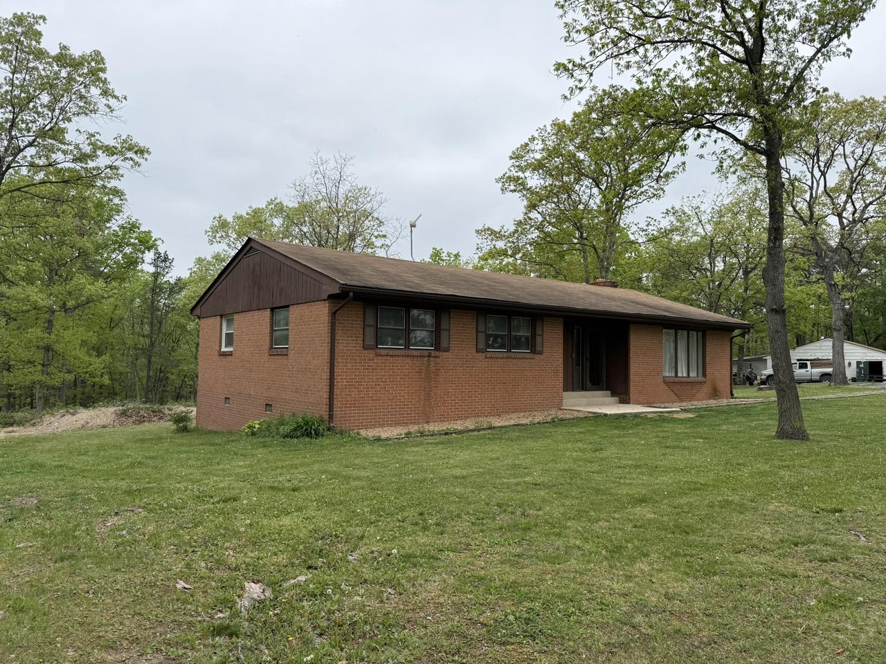 Single-story brick house with a dark gabled roof on a grassy lot surrounded by mature trees.