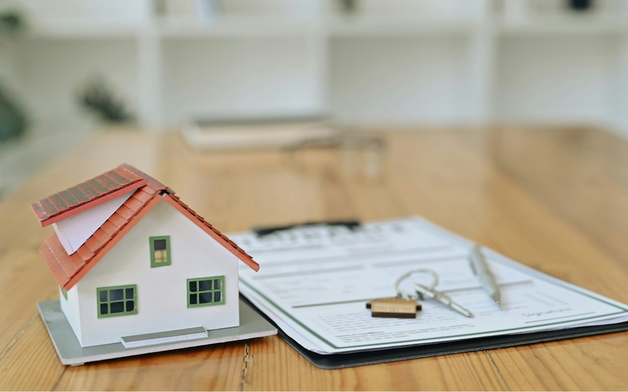 Miniature house model beside real estate paperwork and keys on a wooden table.