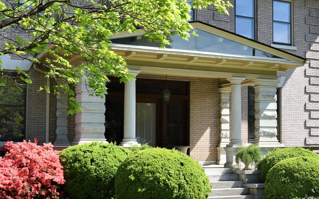 Brick home entrance with a columned portico, front steps, and landscaped shrubs.