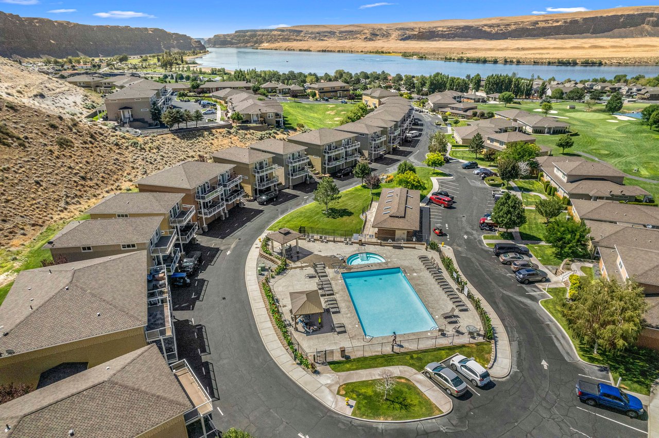 Aerial view of a riverside condo resort with a large pool, parking, and surrounding hills.