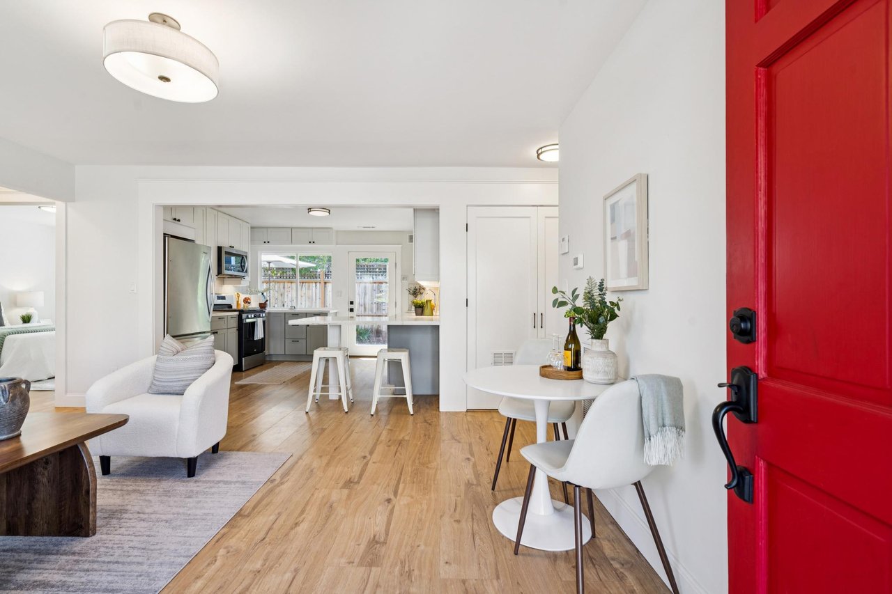 Open-plan living room and kitchen with light wood floors, island seating, and a red entry door.