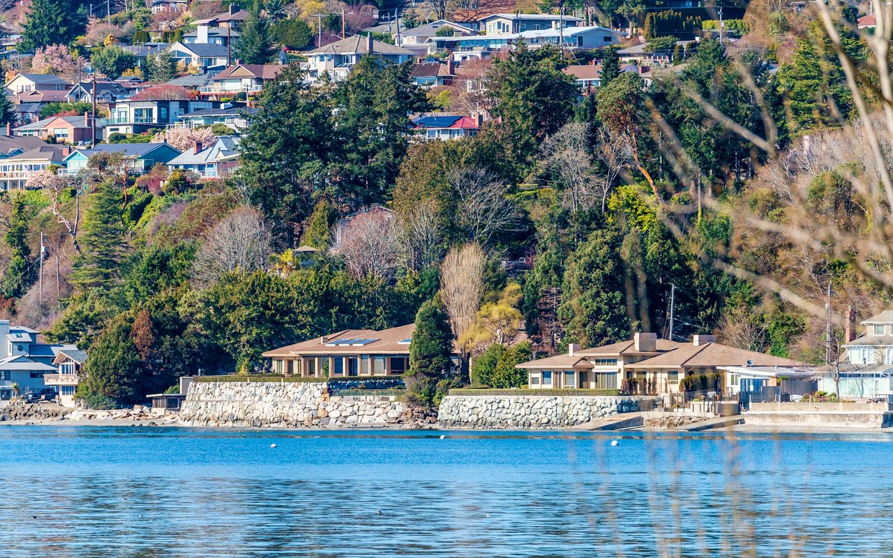 Waterfront homes climb a wooded hillside behind stone seawalls, overlooking blue water.
