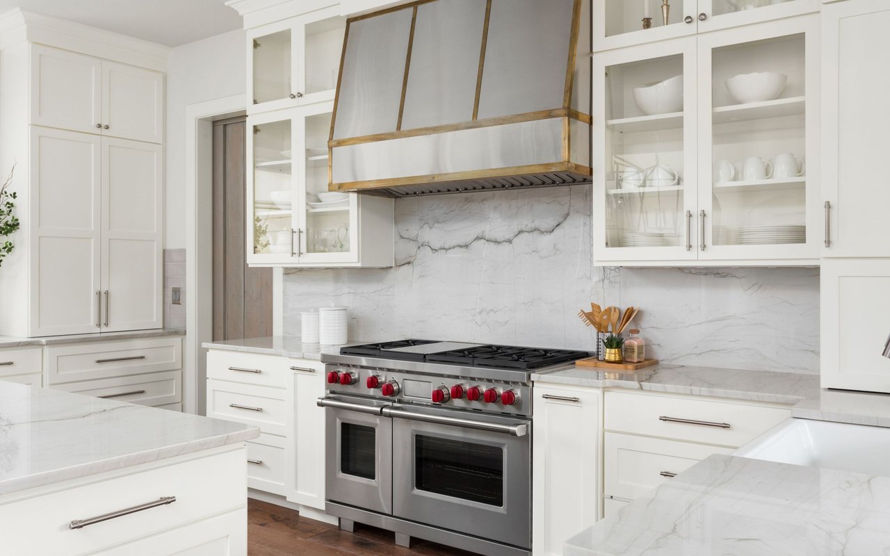 White kitchen with marble surfaces, glass-front cabinets, and a stainless double oven range with red knobs.