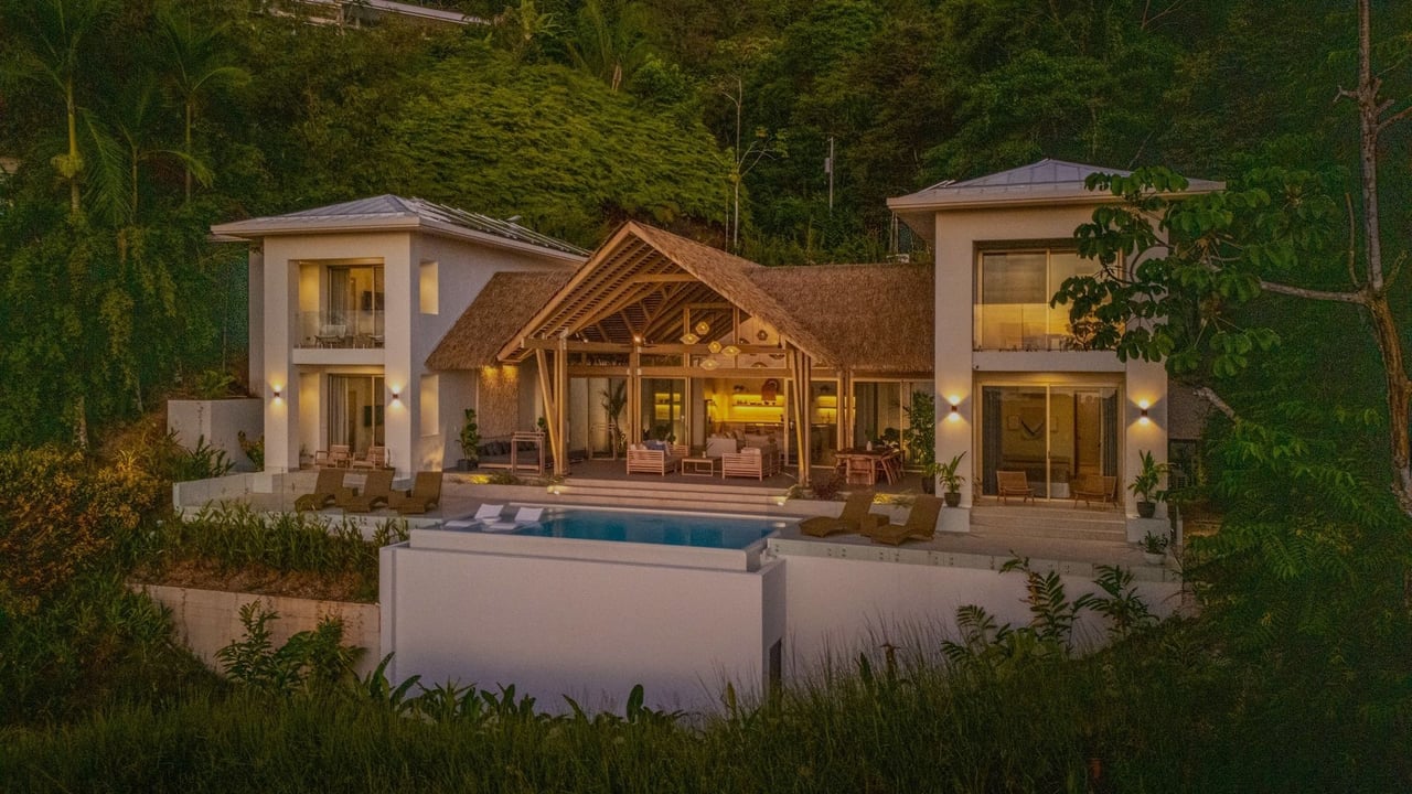 Tropical villa with a thatched central roof, illuminated pool, and dense jungle surroundings.