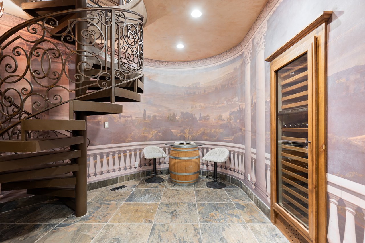 Wine cellar with a wrought-iron spiral staircase, countryside mural, and barrel table between two chairs.