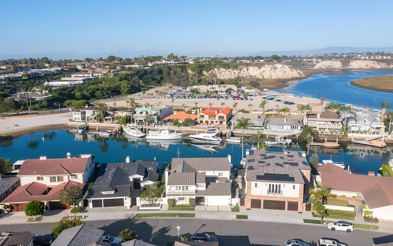 A waterfront neighborhood with modern homes, yachts docked along the canal, and scenic hills in the background.