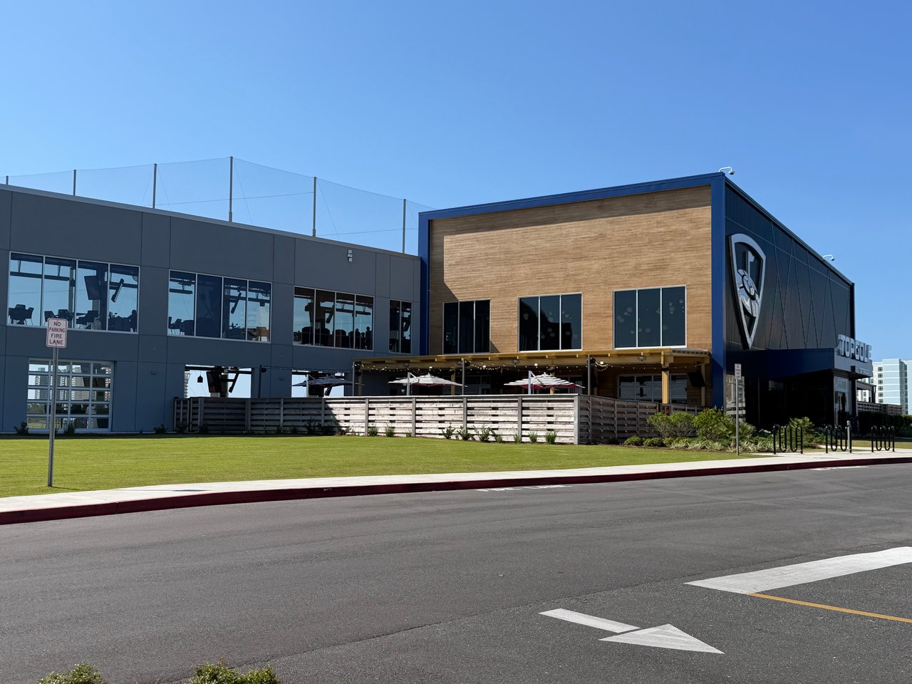 The modern exterior of a multi-story Topgolf sports entertainment complex with a large driving range net and a manicured front lawn.