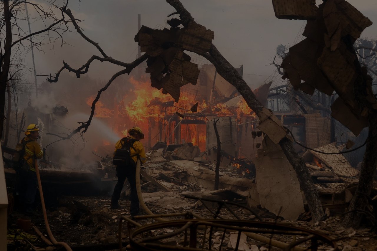  Two firefighters in protective gear, including yellow jackets and helmets, are actively battling a large structure fire. 