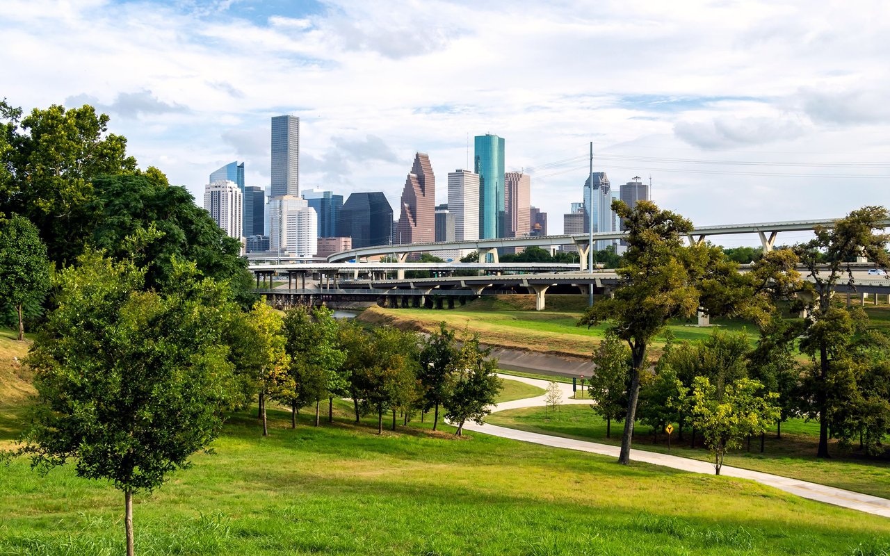 Downtown Houston skyline beyond highway bridges, viewed across a green park and winding path.