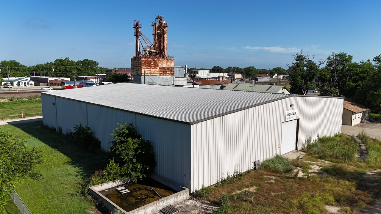 White metal commercial warehouse with a Nutrien Ag Solutions sign and a rusted industrial structure behind it.