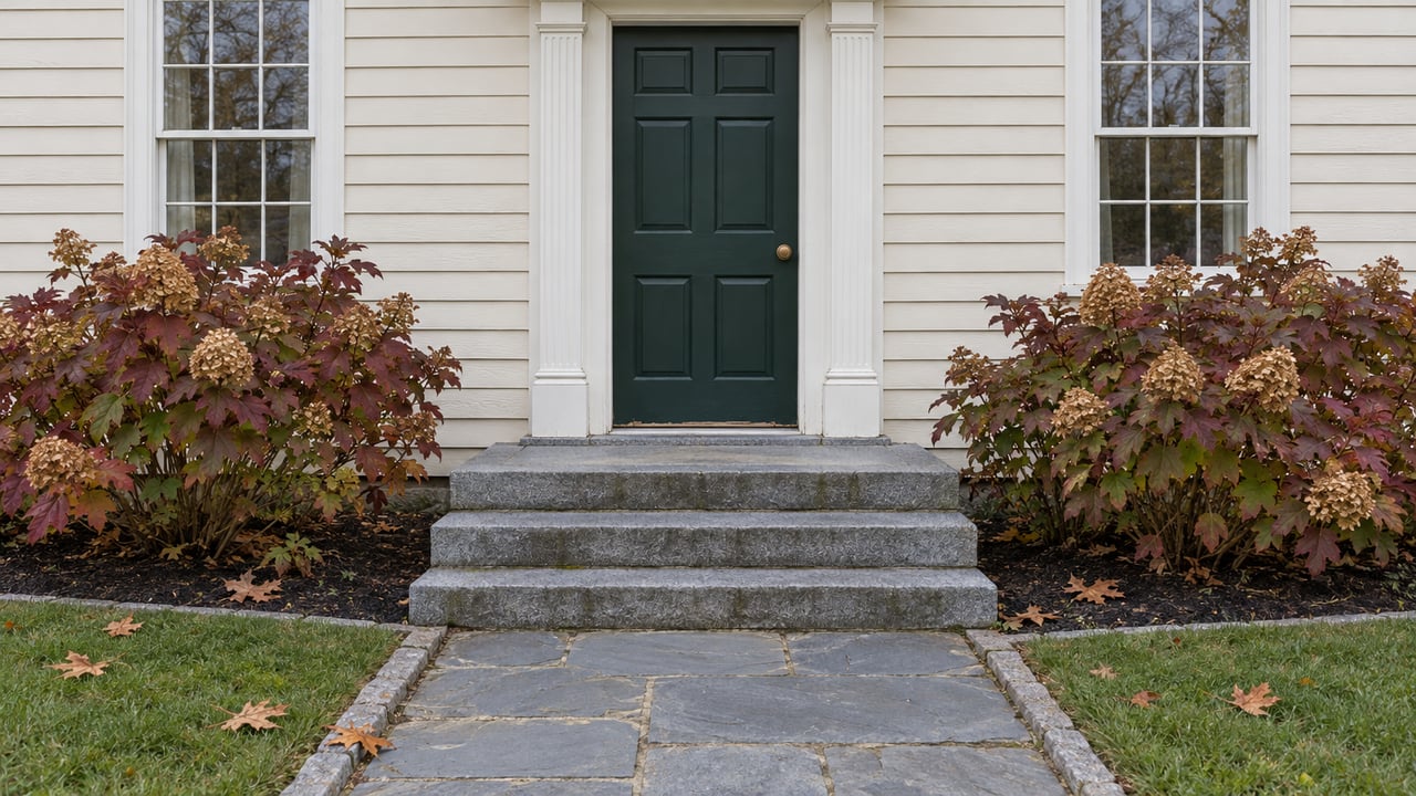 Ivory clapboard entrance with a green paneled door, paired sash windows, granite steps, and burgundy oakleaf hydrangeas beside a bluestone walk.
