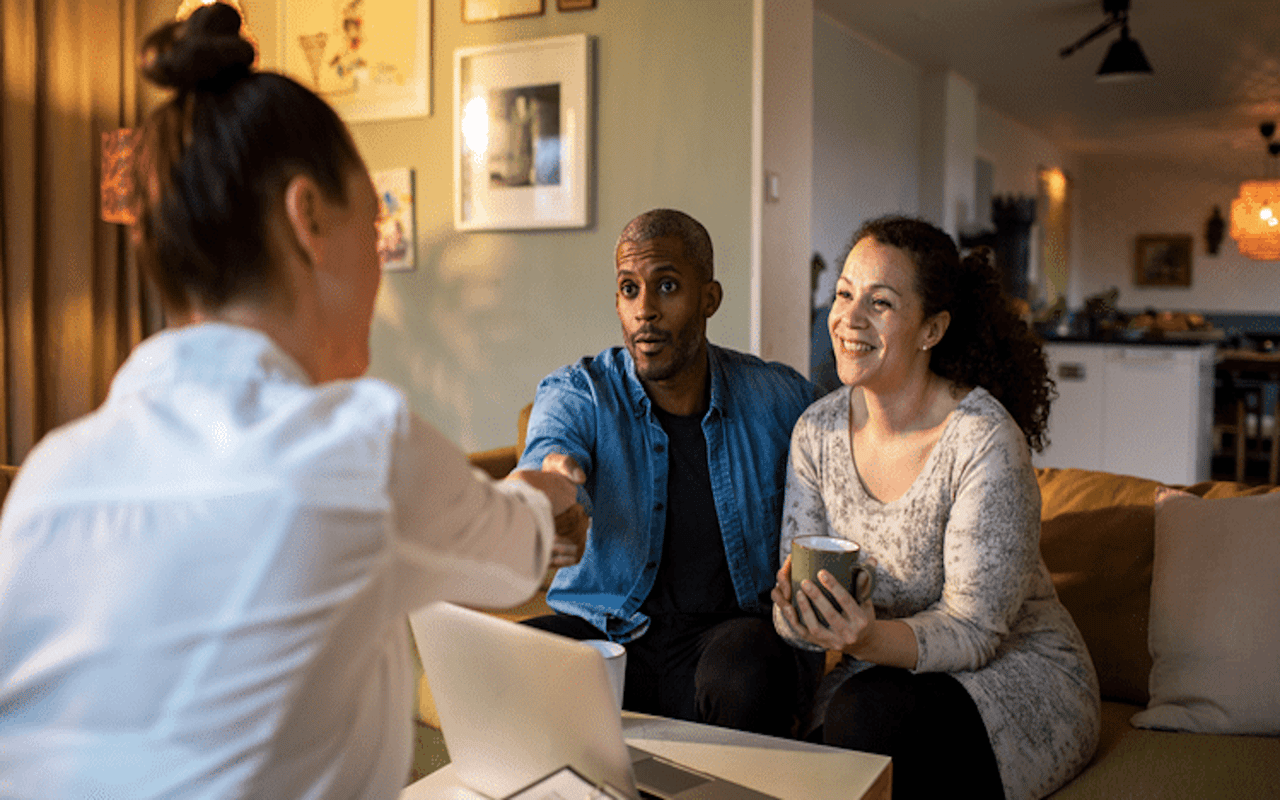 REALTOR® shaking hands with a smiling couple in their living room after securing a home sale.
