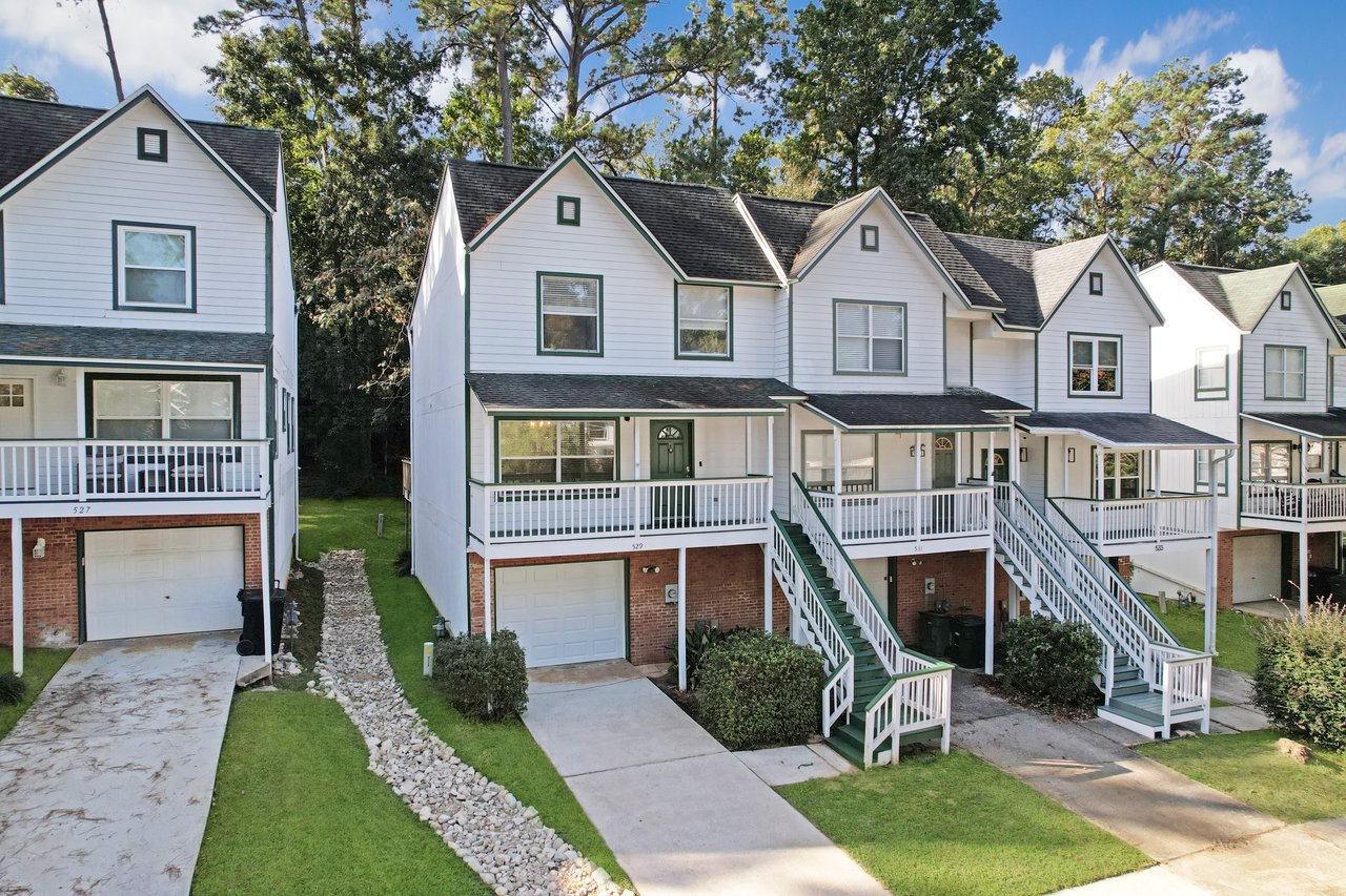 Front exterior view of the townhome at 529 Collinsford Road, Tallahassee, Florida, showing a two-story design with white siding, green trim, and a brick base. The home features a one-car garage, covered front porch with white railings, and green front stairs. A landscaped drainage feature and well-kept lawn enhance the curb appeal.