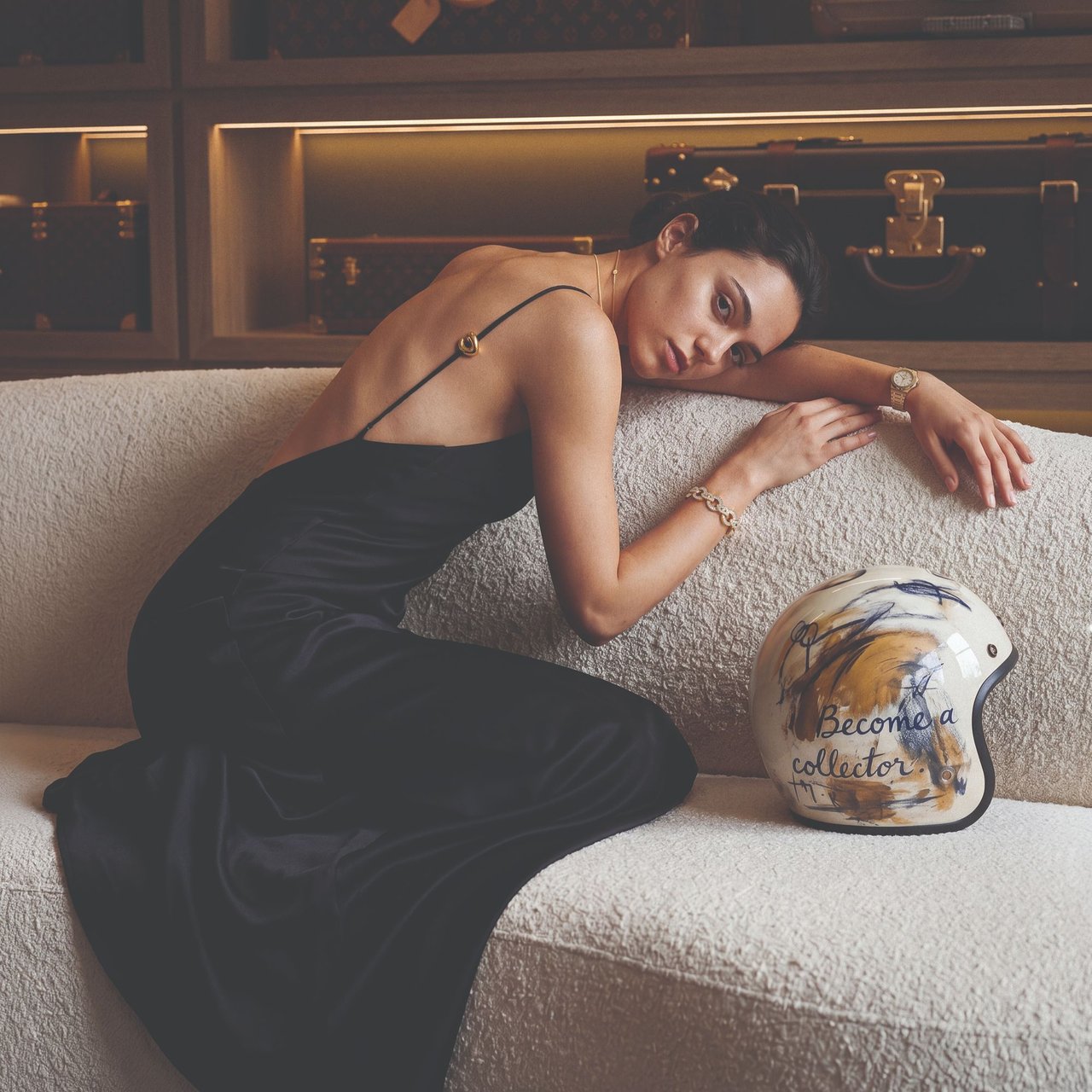 Woman in a black dress reclines on a cream sofa beside a decorated motorcycle helmet.