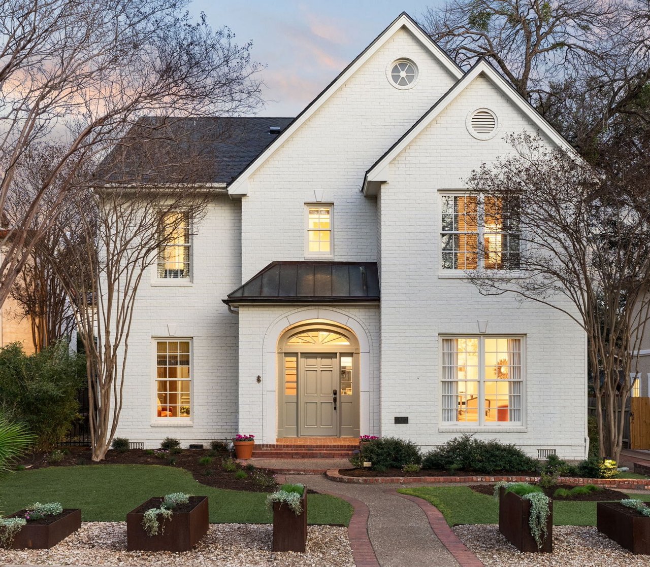 Two-story white brick home with an arched entrance, lit windows, and a landscaped front walkway.