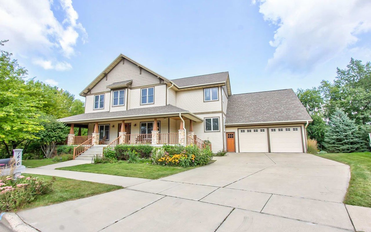 Two-story beige house with a covered porch, stone columns, and an attached two-car garage.