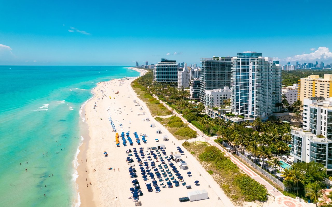 Beachfront condominium towers beside turquoise water and a wide sandy beach lined with umbrellas.