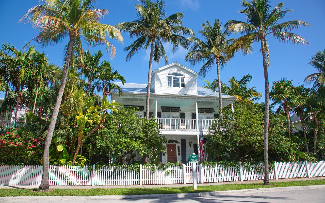 White two-story Key West house with a covered porch, palm trees, and a white picket fence.