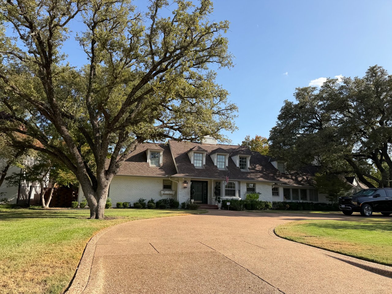 White-brick home with a broad brown roof, mature oaks, and a curved driveway.