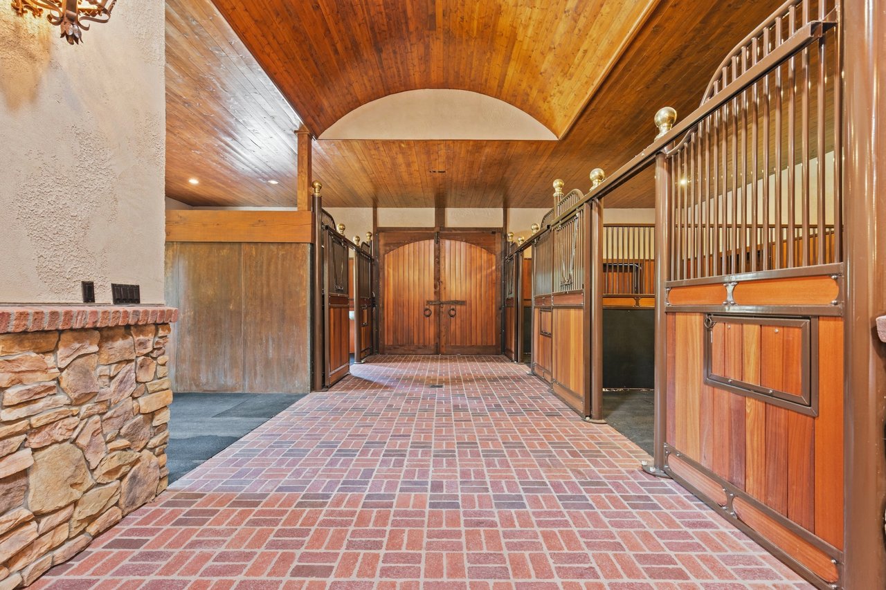 Barn aisle with wooden horse stalls, brick flooring, vaulted wood ceiling, and double doors.
