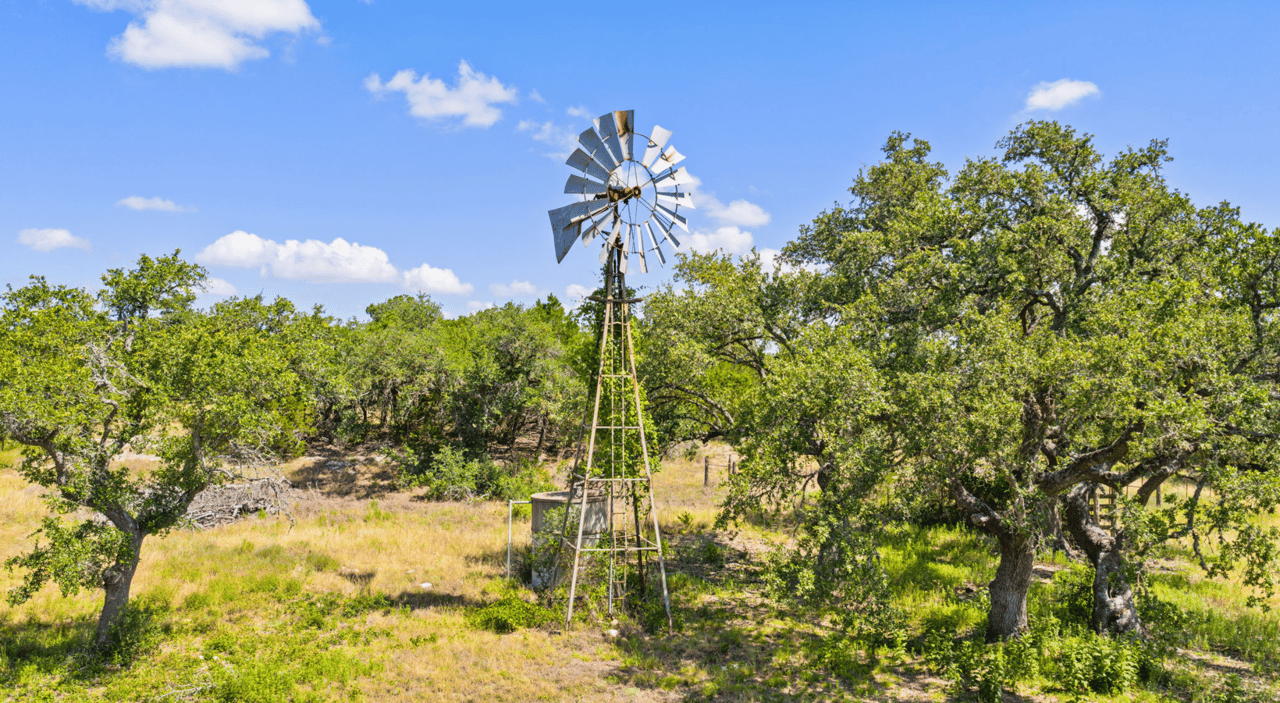 Ranch land with a metal windmill, mature oak trees, and a grassy clearing.
