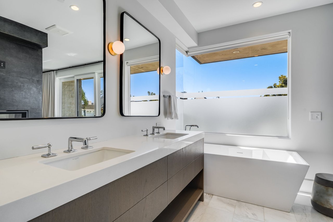 Modern bathroom with a double vanity, black-framed mirrors, and a freestanding tub beside a large window.