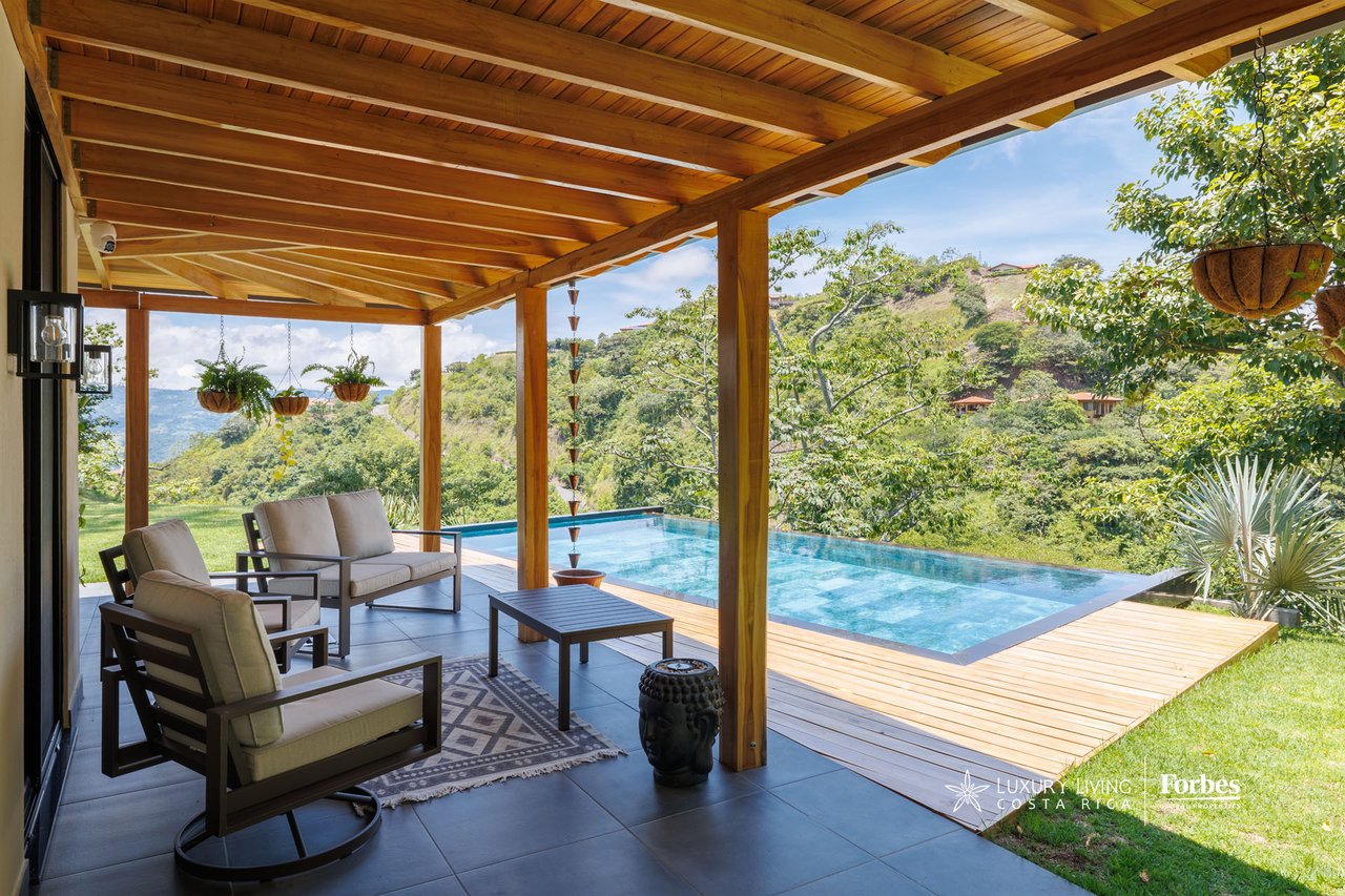 Covered patio with cushioned seating beside an infinity pool overlooking a green valley.