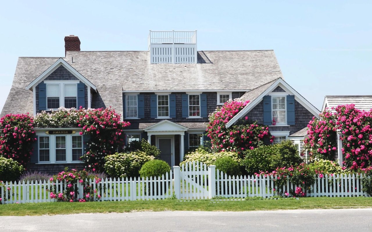 Blue-gray shingle house with a white picket fence and abundant pink flowering shrubs.
