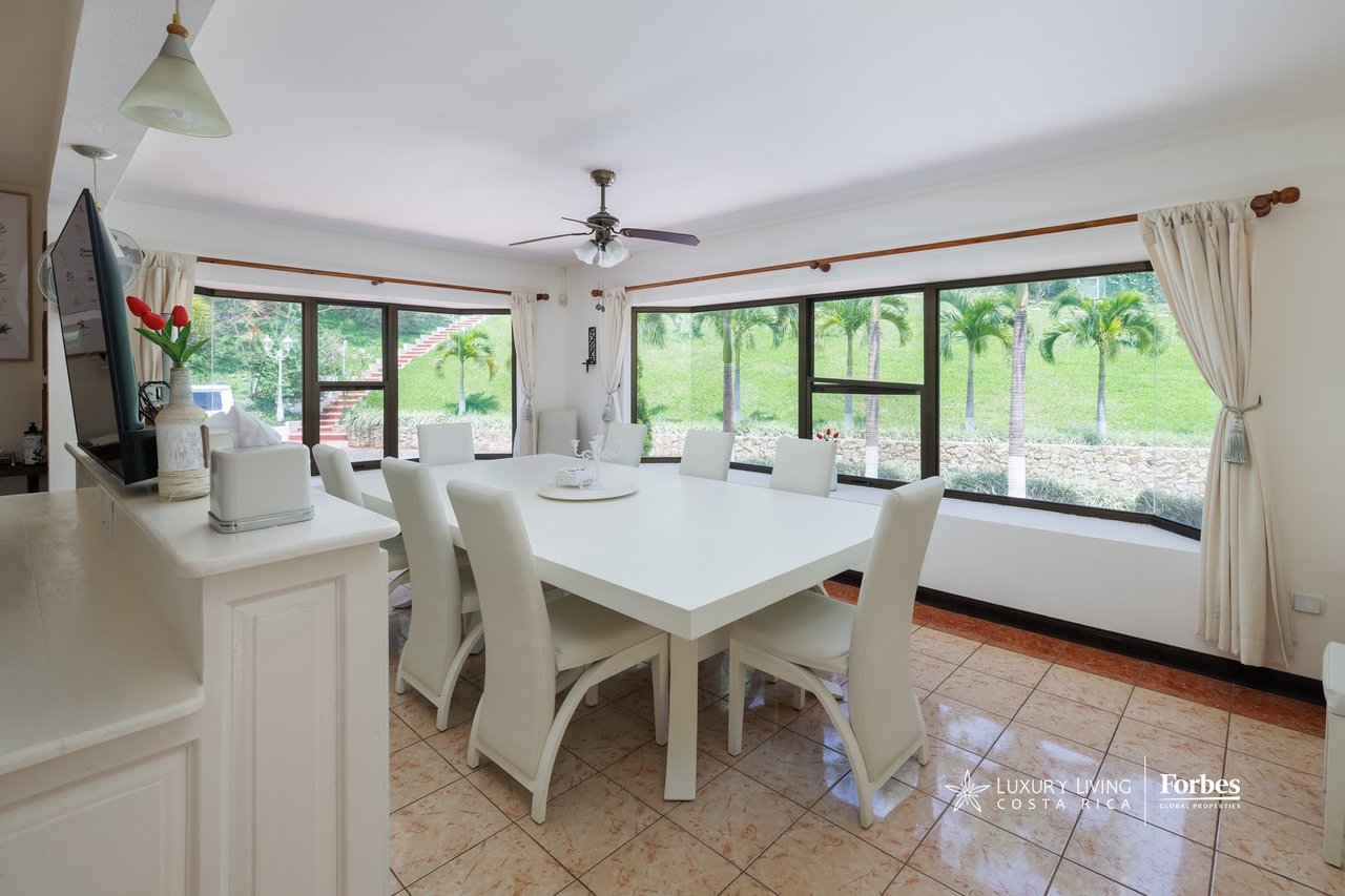 Dining room with a large white table and chairs beside windows overlooking palm trees.