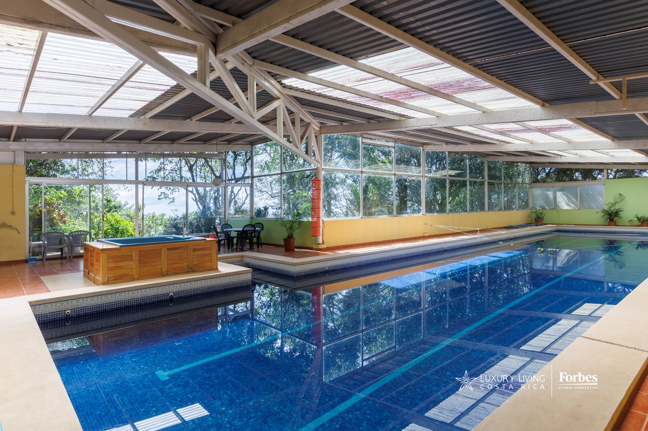 Indoor pool beneath a high metal roof, with a raised wooden hot tub and glass walls.