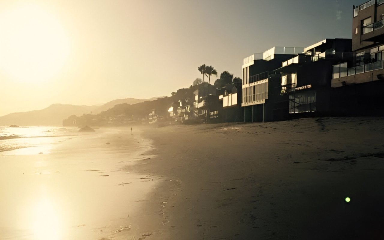 Malibu shoreline at golden hour, with beachfront homes and distant hills.