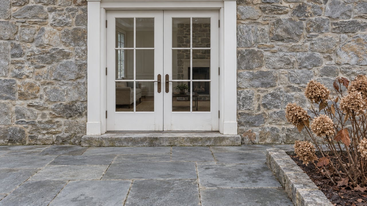 Closed white French doors in a weathered stone wall above a broad stone terrace, with dry oakleaf hydrangea blooms at the corner.