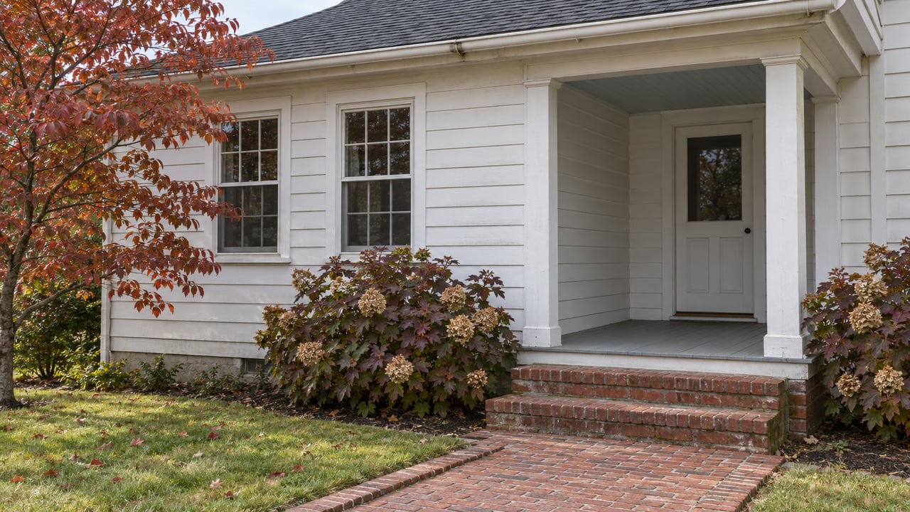 White wood-sided home with multi-pane windows, a covered side porch and worn brick steps beside autumn dogwood and oakleaf hydrangeas.