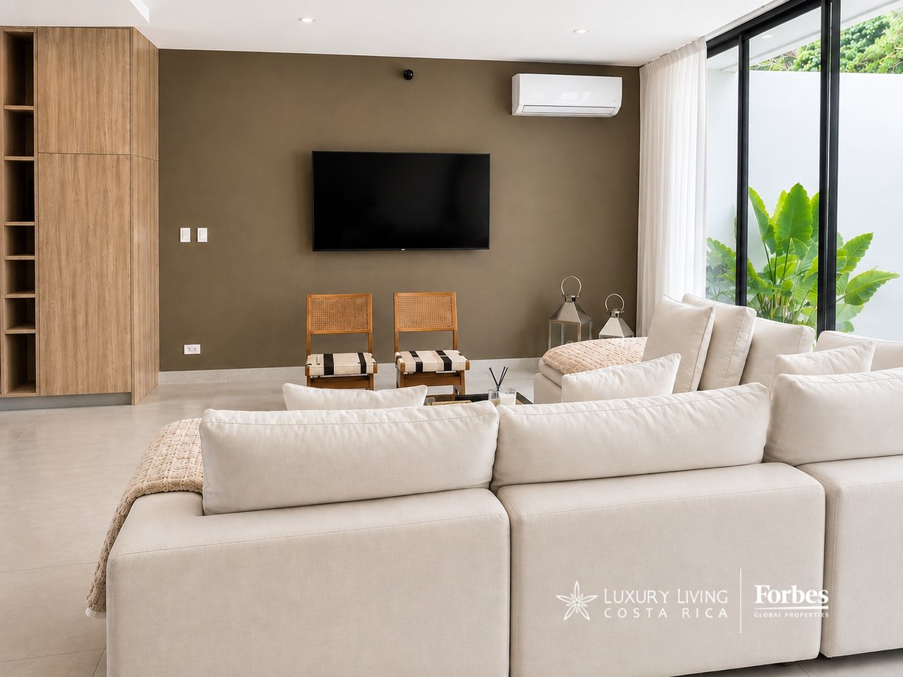 Contemporary living room with cream sectional sofas, cane chairs, and sliding glass doors beside tropical plants.