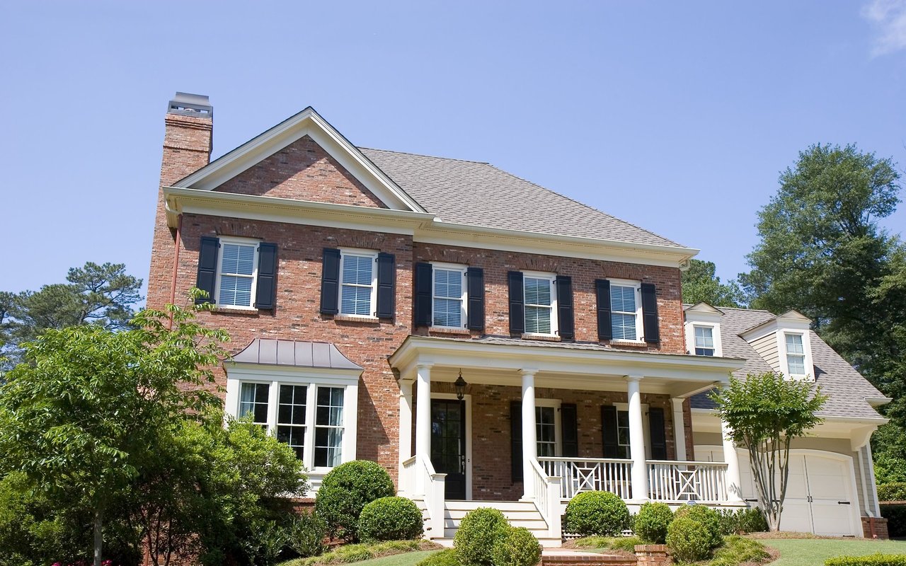 Two-story brick house with a covered porch, dark shutters, and landscaped front yard.