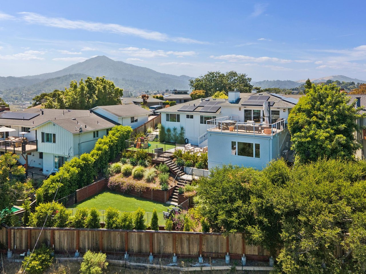 Aerial view of a solar-panelled home with a raised deck and landscaped yard, backed by a mountain.