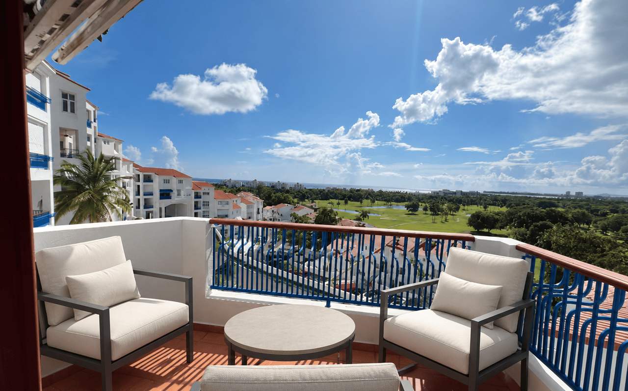 Furnished balcony overlooking a golf course and ocean, with white villas along the left side.