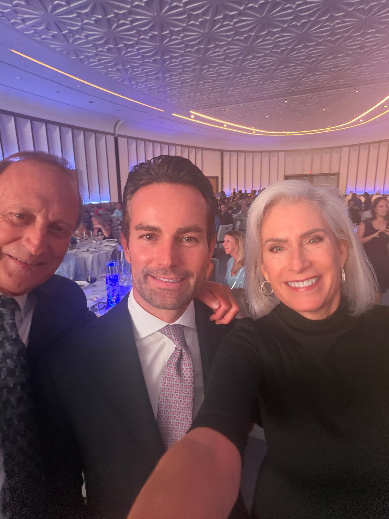 Three adults pose for a selfie at a banquet, with guests seated behind them.