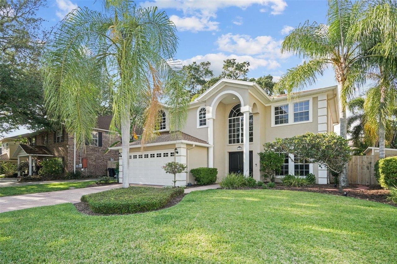 Two-story cream house with an arched entrance, garage, manicured lawn, and palm trees.