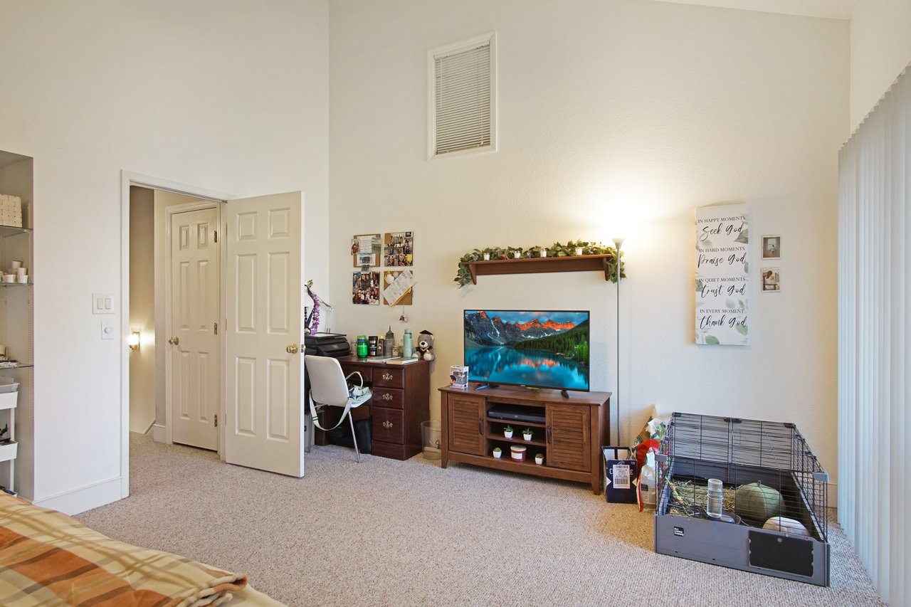 A cozy room with beige walls and carpet, featuring a desk with a chair, a TV on a wooden stand, a pet cage, wall art, and a lighted floor lamp.A cozy room with beige walls and carpet, featuring a desk with a chair, a TV on a wooden stand, a pet cage, wall art, and a lighted floor lamp.