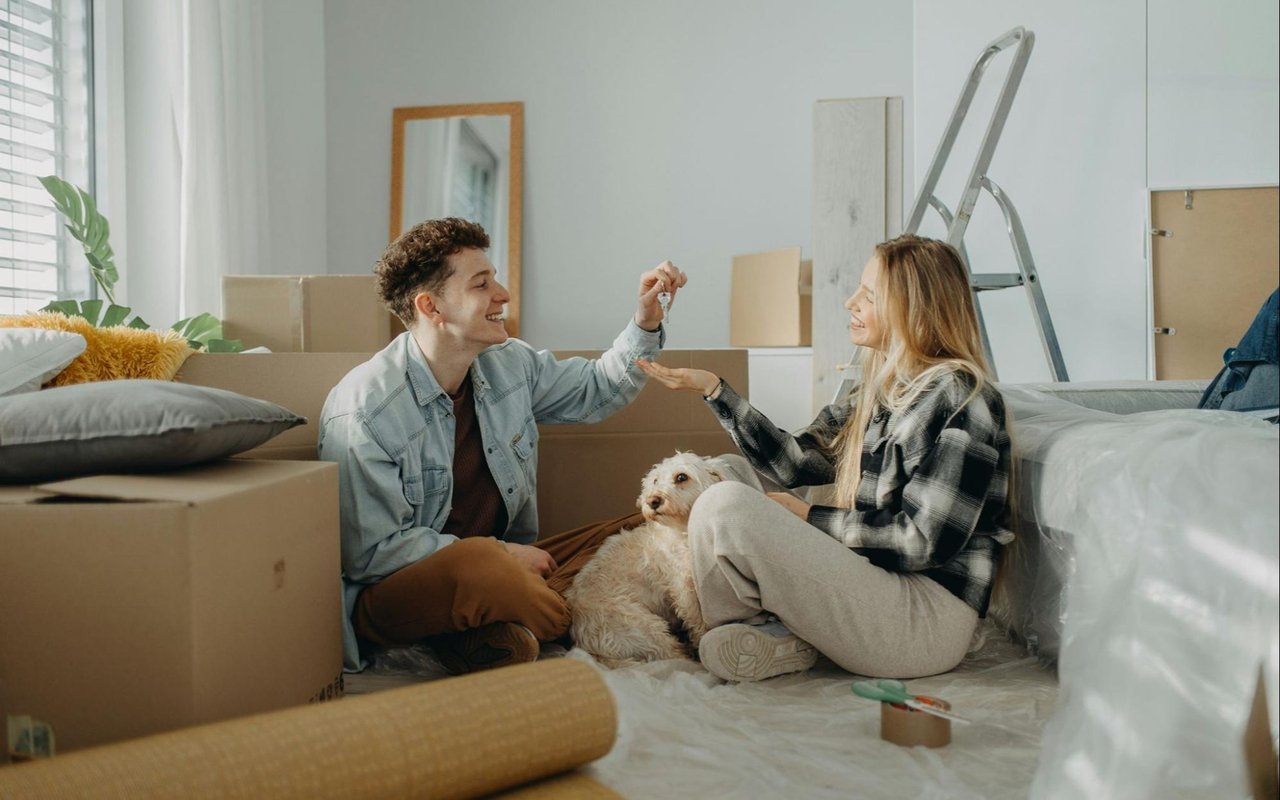 A couple exchanges keys among moving boxes, with their dog beside them.